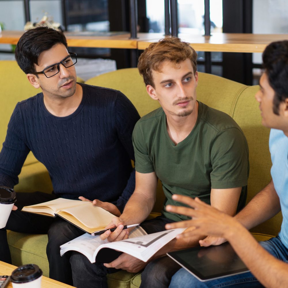 Three serious fellow students discussing homework. Young men sitting on couch at desk. Education and knowledge concept.